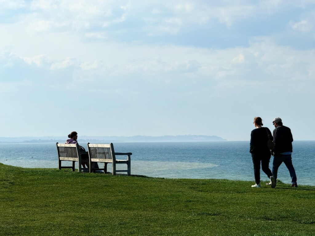 a picture of a bench on a grassy hill over looking the sea with people walking past in whitstable uk