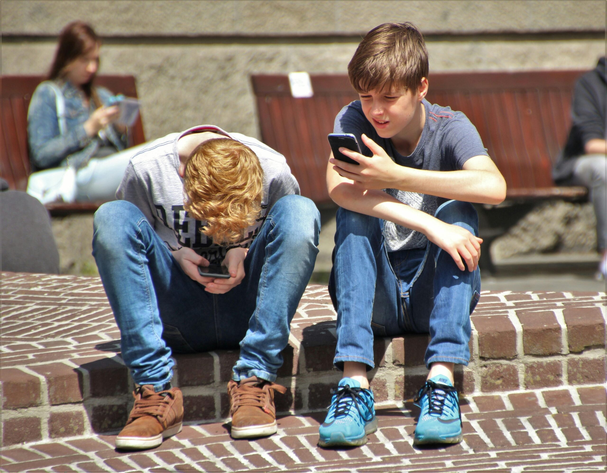 two young teen boys sitting on a tiled step one with his head in between his knnes using a phone one holding a phone in front of his face