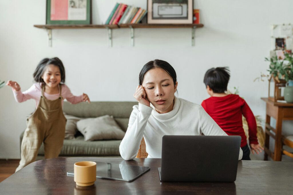 Mum working on a laptop while two children play noisily beside her in a living room