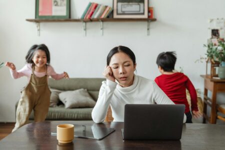 Mum working on a laptop while two children play noisily beside her in a living room
