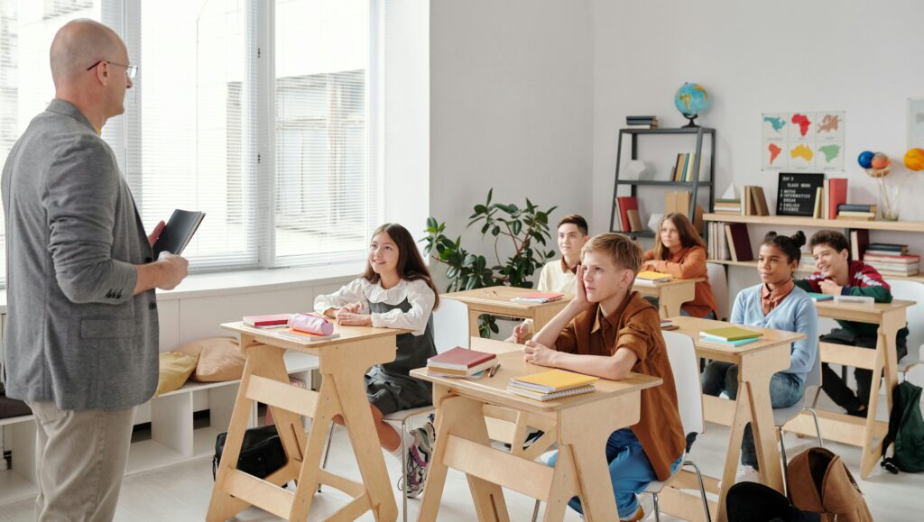 pexels-max-fischer-5212326 students sitting at desks in a classroom listening to their teacher